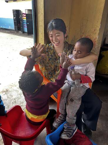 Eine Frau sitzt mit zwei Kindern in einem Raum mit gelben Wänden und einer Tür nach draußen, wahrscheinlich eine Kindertagesstätte oder ein Kindergarten.