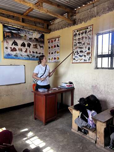 Das Bild zeigt eine Frau, die mit einem langen Stock in der Hand in einem Klassenzimmer steht, mit einem Schreibtisch und einem Stuhl vor ihr, umgeben von Postern und einer Tafel an den Wänden.
