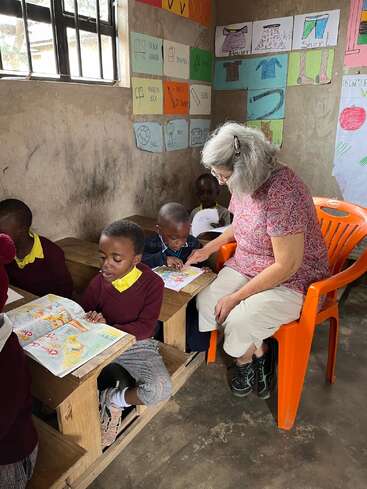 Das Bild zeigt eine Frau, die einer Gruppe von Kindern in einem Klassenzimmer vorliest. Die Kinder sitzen an den Tischen und die Frau sitzt auf einem orangefarbenen Stuhl.