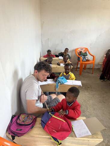 Ein Mann sitzt mit einem Kind an einem Schreibtisch, umgeben von anderen Kindern, in einem Klassenzimmer und hilft wahrscheinlich bei den Schularbeiten. Der Raum ist einfach und konkret.