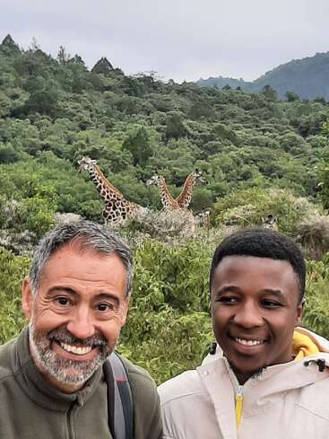 Das Bild zeigt zwei Männer, die vor einer üppigen grünen Landschaft mit Giraffen im Hintergrund für ein Foto posieren und so ein Gefühl von Abenteuer und Kameradschaft vermitteln.