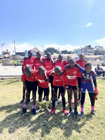 Eine Jugendfußballmannschaft posiert draußen auf einer Wiese und trägt passende rote Trikots. Sie stehen in zwei Reihen zusammen und lächeln unter dem strahlend blauen Himmel.