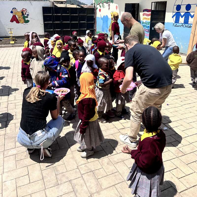Eine Gruppe kleiner Kinder in Uniformen versammelt sich draußen und unterhält sich freudig mit Erwachsenen, die in der Nähe eines bunten, mit Wandmalereien verzierten Gebäudes unter einem strahlenden Himmel Essen oder Leckereien verteilen.