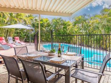 The image depicts a serene outdoor dining area with a pool, surrounded by palm trees, featuring a table set for two with a bottle of wine and a covered patio with lounge chairs.