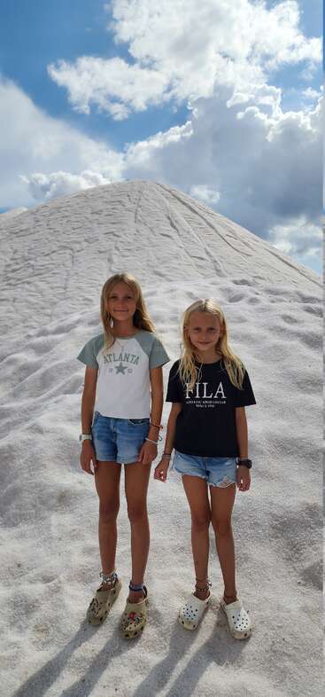 Two young girls stand on a large white mound, possibly salt or sand, under a partly cloudy sky, wearing shorts, t-shirts, and sandals, smiling.