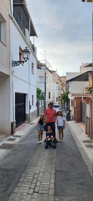 A man with a stroller and three children walk down a narrow street surrounded by houses, lamp posts, and parked vehicles under a partly cloudy sky.