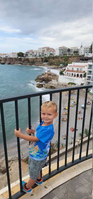 A young boy smiles while holding a black railing, standing above a scenic beach with sunbathers, ocean waves, rocky cliffs, and white buildings under a cloudy sky.