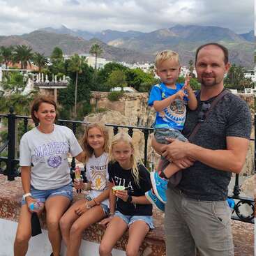 A family of five poses outdoors with mountains and greenery in the background. The children enjoy ice cream, and everyone looks relaxed and happy, enjoying vacation.