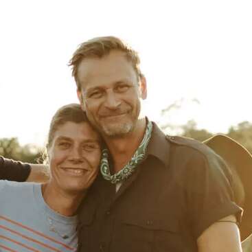 Una pareja sonriente posa muy unida al aire libre, con aspecto feliz y relajado. La luz del sol crea un cálido resplandor dorado en el fondo, lo que realza el ambiente alegre.