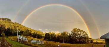 Un double arc-en-ciel traverse un paysage rural composé d'arbres verts, de champs ouverts, d'un chemin de terre, de clôtures en bois et d'un hangar à toit métallique, le tout sous un ciel nuageux.