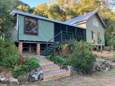 Cette image montre une charmante maison verte en bois sur pilotis, avec un porche couvert, entourée de jardins luxuriants, de marches en pierre et de grands arbres en arrière-plan.