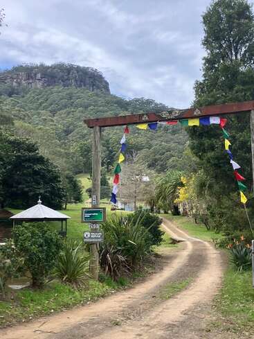 Un paysage de montagne serein avec un chemin de terre qui mène à une arche en bois décorée. Des drapeaux de prière colorés sont suspendus ; des arbres, un pavillon et des panneaux indiquant un refuge pour animaux sauvages sont visibles.