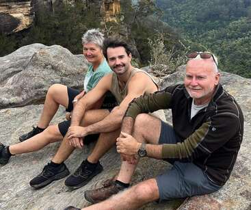 Three people sit together on a rocky ledge, smiling and enjoying a scenic outdoor view, surrounded by greenery and distant cliffs, dressed in casual hiking clothes.