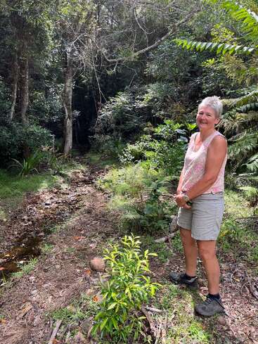 Uma mulher sorridente está em uma clareira de uma floresta exuberante ao lado de um pequeno riacho rochoso, cercada por plantas verdes, árvores e luz do sol filtrada pelas folhas.