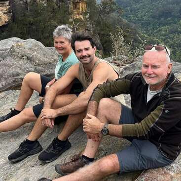 Três pessoas sentadas juntas em uma borda rochosa, sorrindo e desfrutando de uma vista panorâmica ao ar livre, cercadas por vegetação e penhascos distantes, vestidas com roupas casuais de caminhada.