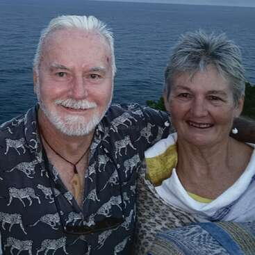 Un couple âgé, souriant et enlacé, pose ensemble en plein air près de l'océan. L'arrière-plan montre une eau et un ciel calmes, créant un moment serein et heureux.