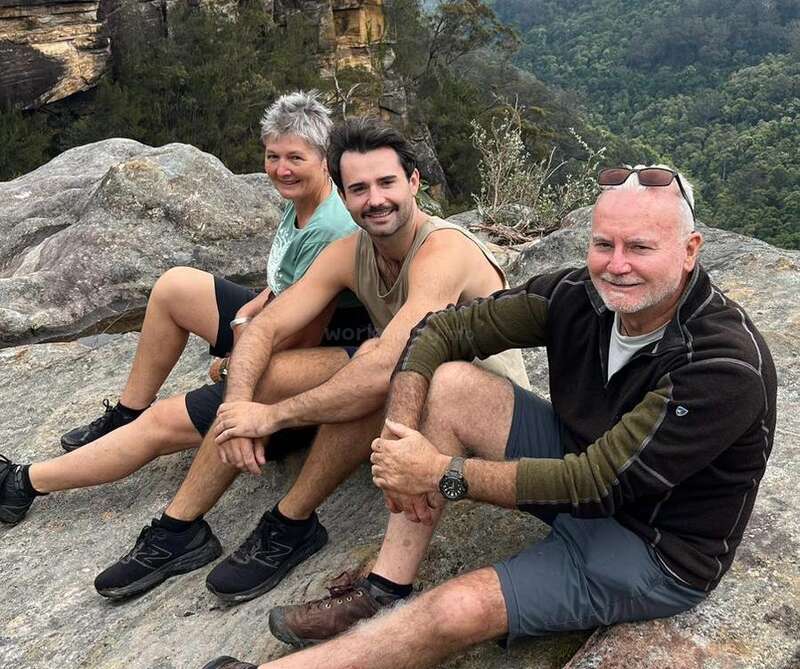 Trois personnes sont assises ensemble sur une corniche rocheuse, souriant et profitant d'une vue panoramique en plein air, entourées de verdure et de falaises lointaines, vêtues de vêtements de randonnée décontractés.