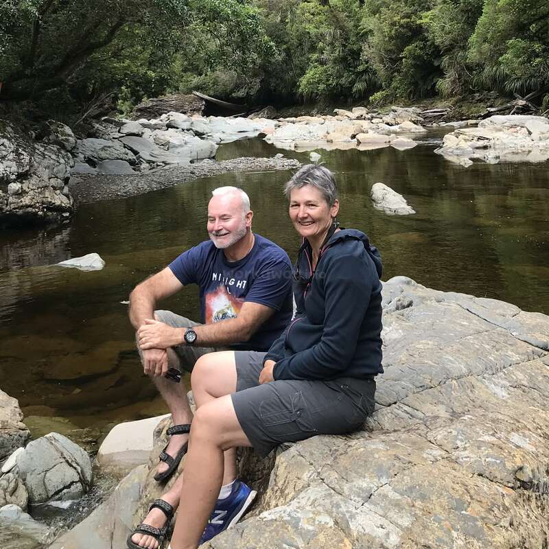 Um casal sorridente senta-se em grandes rochas ao lado de um rio tranquilo e rochoso. Cercados por uma floresta verdejante, eles curtem a natureza, relaxam e aproveitam a companhia um do outro.