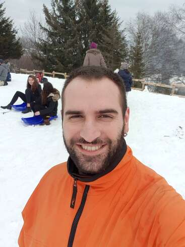 Um homem sorridente com uma jaqueta laranja tira uma selfie em uma paisagem de neve. As pessoas atrás dele estão sentadas em trenós, com árvores ao fundo.