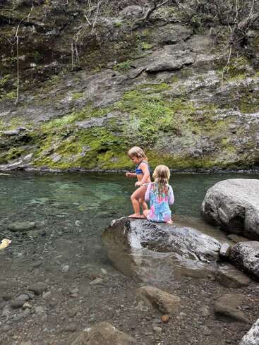 Zwei kleine Kinder sitzen auf einem großen Stein neben einem klaren, flachen Bach, umgeben von moosbewachsenen Felsen und viel Grün, und genießen gemeinsam einen ruhigen Moment in der Natur.