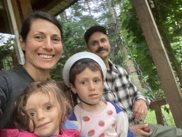 Una familia de cuatro miembros posa sonriente al aire libre. La mujer está delante, con dos niños, uno de ellos con casco. Un hombre está de pie detrás de ellos. Los árboles les rodean.