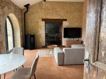 Ein gemütliches Wohnzimmer mit Steinwänden, einem runden Esstisch, grauen Stühlen, einem Fernseher, einem Sofa, einem Holzofen, einem Bogenfenster und einem bunten Teppich.