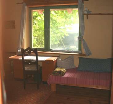 This cozy room features a window with green trim, wooden desk, cushioned chair, daybed with colorful pillows, and leafy tree view outside, bathed in daylight.