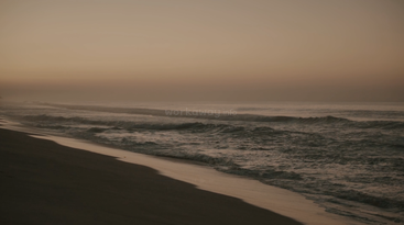 A serene beach scene at sunset, with gentle waves rolling onto the shore. The sky glows softly in orange hues, creating a peaceful, calming atmosphere.