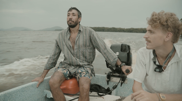 Two men are sitting in a small motorboat on a body of water. One is steering, while the other looks thoughtfully ahead. The scenery is calm.