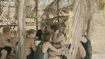 A group of six people sit under a rustic palm-leaf shelter, gathered around a wooden table, talking and sharing a meal in a tropical setting.