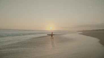A lone surfer walks along a serene beach at sunset, carrying a surfboard, with gentle waves lapping the shoreline and the sky painted golden orange.