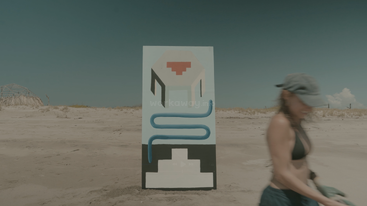 A rectangular artwork with abstract design stands on a sandy beach under a clear sky. A woman in a hat and black bikini walks by in front.