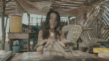 A woman with long hair shapes clay with her hands in a rustic, sunlit outdoor workshop, surrounded by tools, water containers, and colorful fabric decorations.