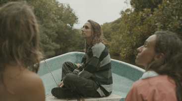 Three people are on a small boat surrounded by lush greenery. The person in the center, wearing a striped shirt, looks thoughtfully upward. It's a peaceful moment.