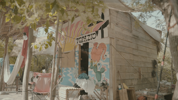 A rustic wooden hut with colorful murals, including a sunrise and rooster, surrounded by trees, hammocks, and outdoor furniture, creating a relaxed, tropical atmosphere.