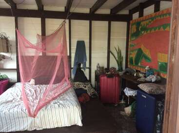 A cozy room with a bed covered by a pink mosquito net, luggage, a colorful wall painting, plant, and assorted belongings scattered on a desk and floor.