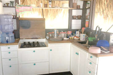 A rustic kitchen with white cabinets, a gas stove, water dispenser, dishes, utensils, food items, open shelves, and a thatched roof visible through the window.