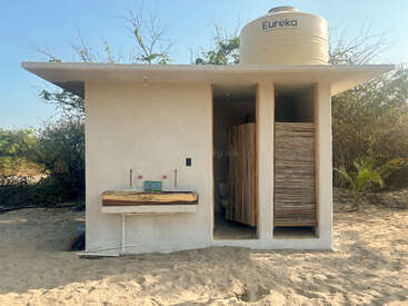 Dieses Bild zeigt ein einfaches Bad im Freien mit zwei Kabinen, einem Wassertank mit der Aufschrift "Eureka", einem rustikalen Waschbecken aus Holz und einer sandigen Umgebung mit spärlicher Vegetation.