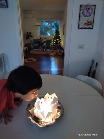 A child leans over a table, gazing at illuminated paper houses. In the background, a lit Christmas tree and cozy living room create a festive atmosphere.