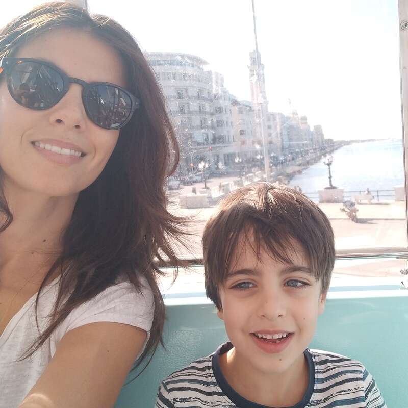 A woman wearing sunglasses and a boy are smiling for a selfie. They are outside, near a waterfront with historic buildings and bright sunlight.
