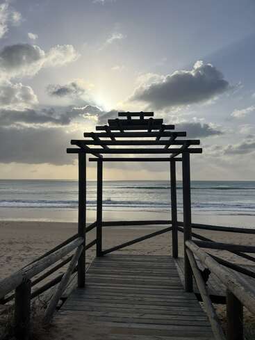 Une promenade en bois avec une pergola mène à une plage de sable. Le soleil se couche derrière des nuages spectaculaires, projetant des ombres et des reflets lumineux sur les vagues calmes de l'océan.
