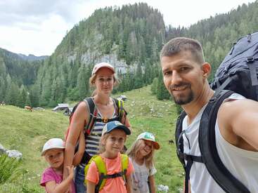 The image depicts a family of five posing for a selfie in a picturesque grassy field, surrounded by trees and mountains, with a cloudy sky above.
