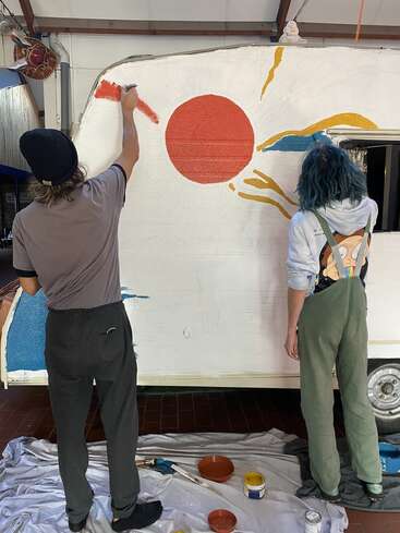 Zwei Personen malen ein Wandgemälde auf einen weißen Camper. Das Kunstwerk zeigt eine große rote Sonne, gelbe und blaue Strahlen und verschiedene Farbdosen auf dem Boden.