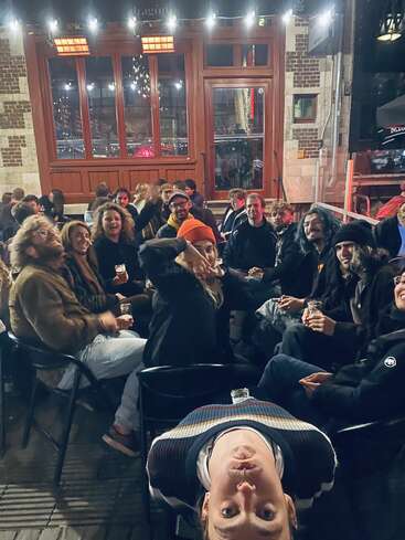 Eine lebhafte Gruppe von Freunden sitzt nachts vor einem Café und lächelt. Eine Person posiert kopfüber im Vordergrund und verleiht ihr spielerische Energie.