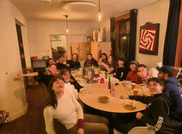 Eine Gruppe von Freunden versammelt sich um einen Tisch und genießt eine gemeinsame Mahlzeit in einem gemütlichen, warm beleuchteten Raum, wo sie lachen, Suppe, Getränke und fröhliche Gespräche miteinander teilen.