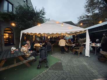 Die Menschen versammeln sich unter einem weißen Zeltdach und genießen das Essen und die Gespräche. Lichter und Bäume im Freien schaffen eine gemütliche Atmosphäre. Tische, Stühle und ein Kinderwagen sind zu sehen.