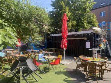 Dieses Außencafé bietet vielseitige Sitzgelegenheiten, bunte Stühle, runde Tische, Lichterketten, einen großen roten Schirm, üppige grüne Bäume und urbane Kunst an den Wänden.