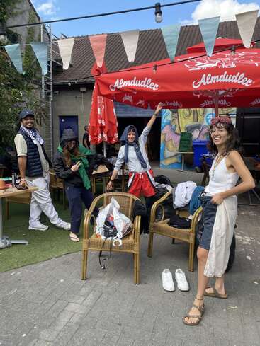 Vier Freunde posieren fröhlich unter einem roten Almdudler-Regenschirm im Freien. Darüber hängen bunte Fahnen. Zwei Korbstühle, eine Kamera, Schuhe und Taschen vervollständigen die lebhafte Szene.