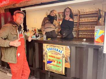 Drei fröhliche Menschen posieren an einem Bartresen im Freien, zwei Frauen hinter der Theke lächeln, ein Mann in roter Latzhose zeigt den Daumen nach oben, festliche Stimmung, Gläser sind zu sehen.