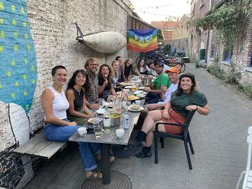 Eine Gruppe von Freunden isst gemeinsam an einem langen Tisch im Freien. An der Wand hängen eine bunte "PACE"-Friedensflagge und ein gemaltes Wandgemälde.
